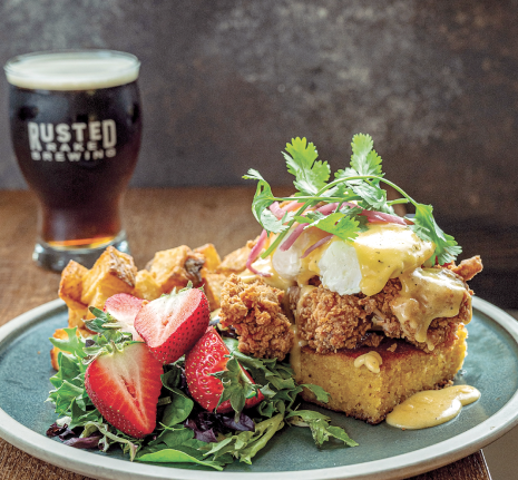 Fried Chicken Benny with house-made cornbread, crispy fried chicken, poached eggs, hollandaise, pickled onions, and cilantro, served with potatoes, greens, and fruit at Rusted Rake Brewing